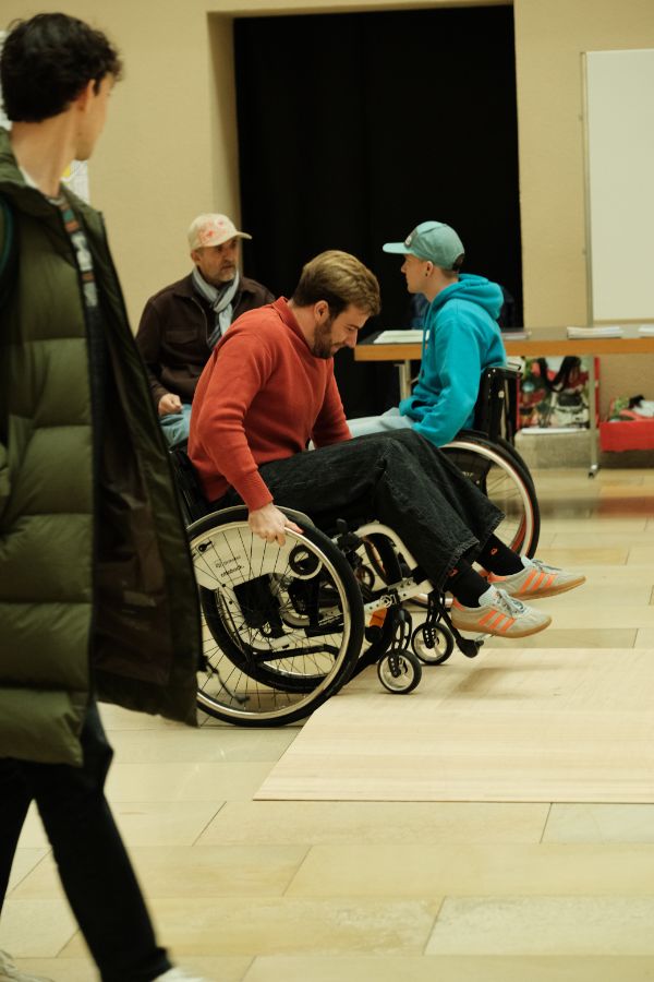 Rollstuhlsimulation: Eine Person im Rollstuhl bewegt sich aktiv vorwärts, während andere Personen im Hintergrund stehen oder sitzen.